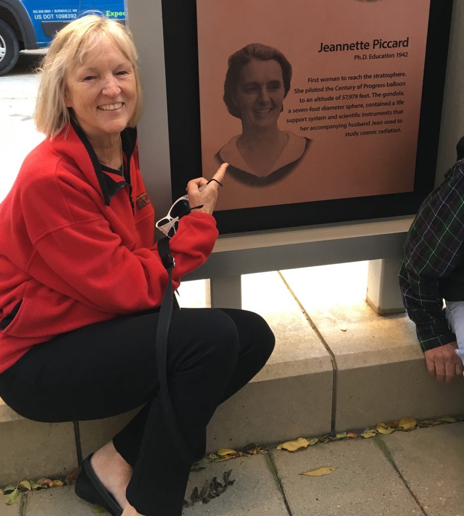 Color photo of Jane Piccard sitting on a ledge pointing to her grandmother's photo on a display panel