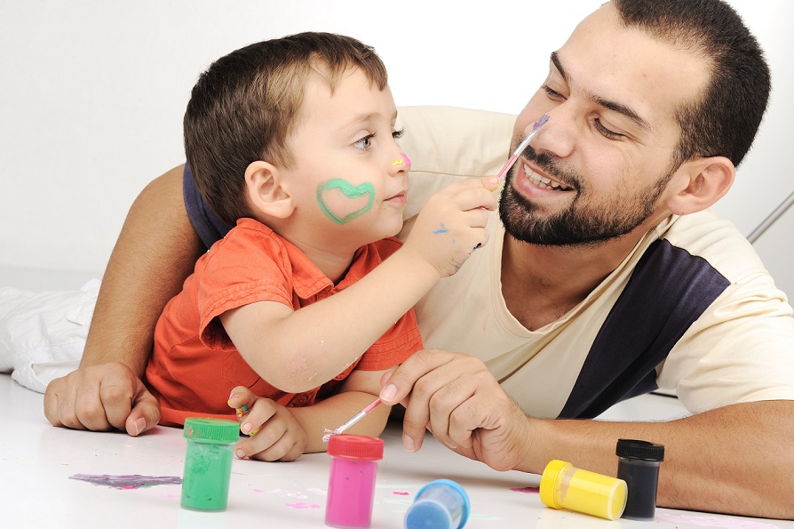 A toddler and adult male caregiver play with paint as the toddler paints a spot on the man's nose.