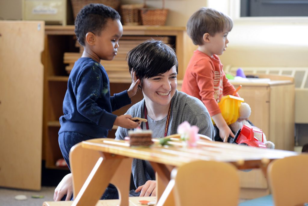A young woman working in a daycare smiles as she interacts with toddlers and a boy touches her hair