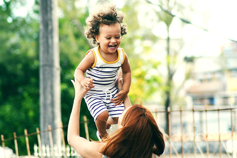 Mother lifts child into the air outside with trees and a fence surrounding them. 