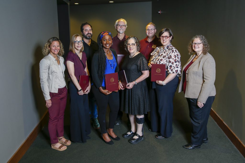 A group of nine people stands together indoors, holding certificates. They are smiling, dressed in business casual and formal attire, with a plain background behind them. Mary R. Georing is in the front row, third from the right. 